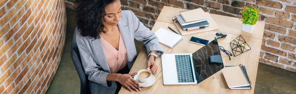 Panoramaaufnahme einer afrikanisch-amerikanischen Geschäftsfrau mit Kaffeetasse und Laptop am Schreibtisch  - Foto, Bild