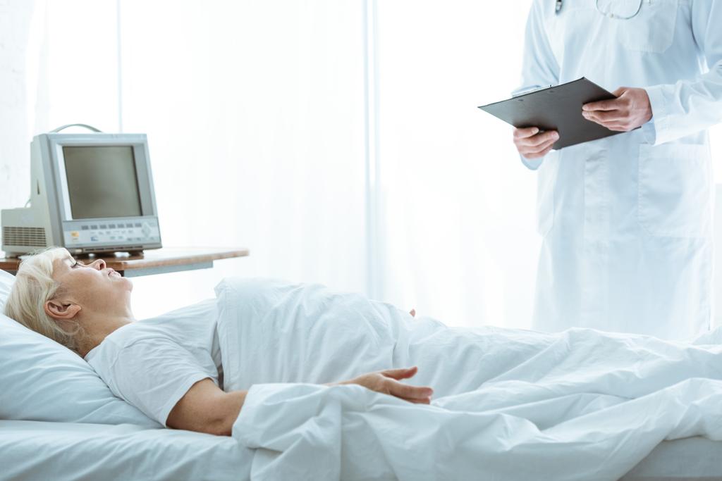 vista ritagliata del medico con appunti e malata donna anziana sdraiata sul letto
 - Foto, immagini