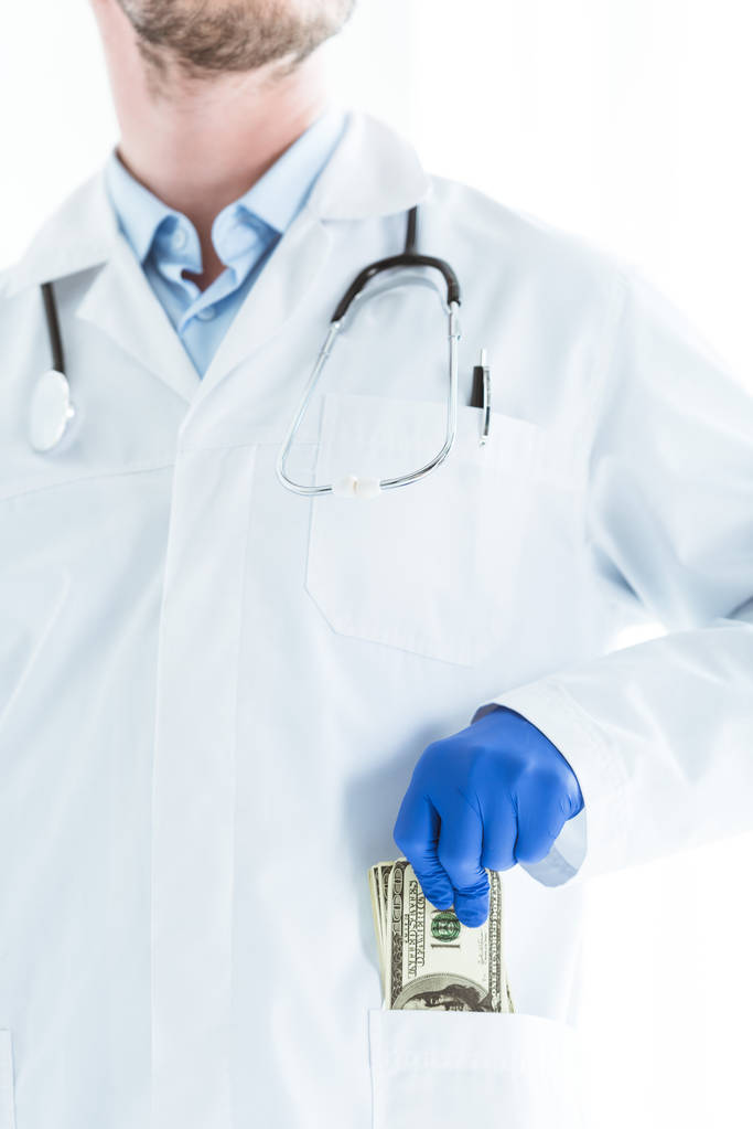 gedeeltelijke weergave van de arts in witte vacht putting dollar bankbiljetten in zak geïsoleerd op wit - Foto, afbeelding