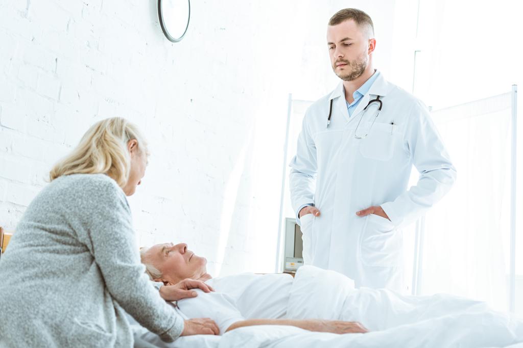 vakava lääkäri seisoo kädet pockers lähellä sairas potilas ja vanhempi nainen klinikalla
 - Valokuva, kuva
