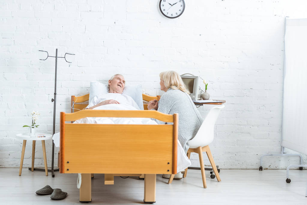 huolestunut vanhempi nainen istuu lähellä sairas husbend ja pitää kätensä sairaalassa
 - Valokuva, kuva