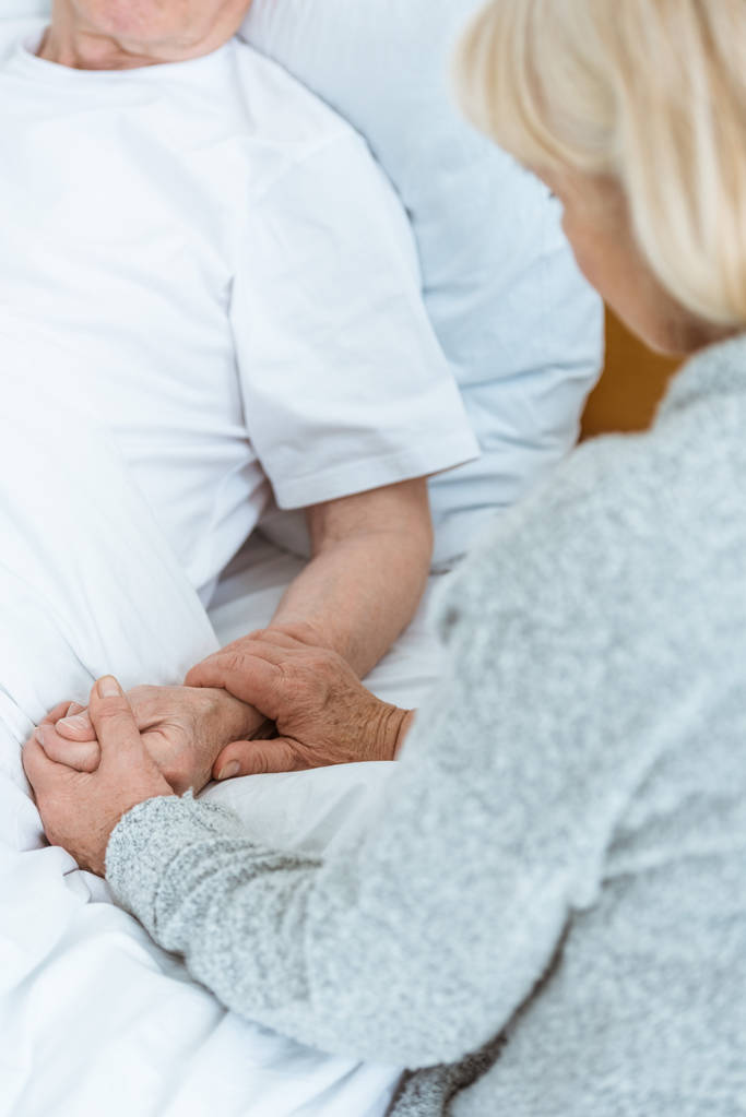 oříznutý pohled na starší ženu držící ruce s nemocným manželem v nemocnici - Fotografie, Obrázek