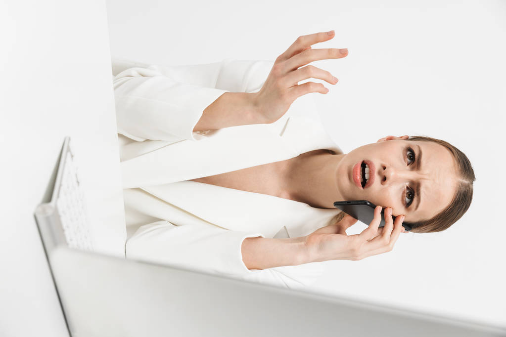 Mujer disgustada sentada aislada sobre fondo de pared blanca usando ordenador PC hablando por teléfono móvil
. - Foto, imagen