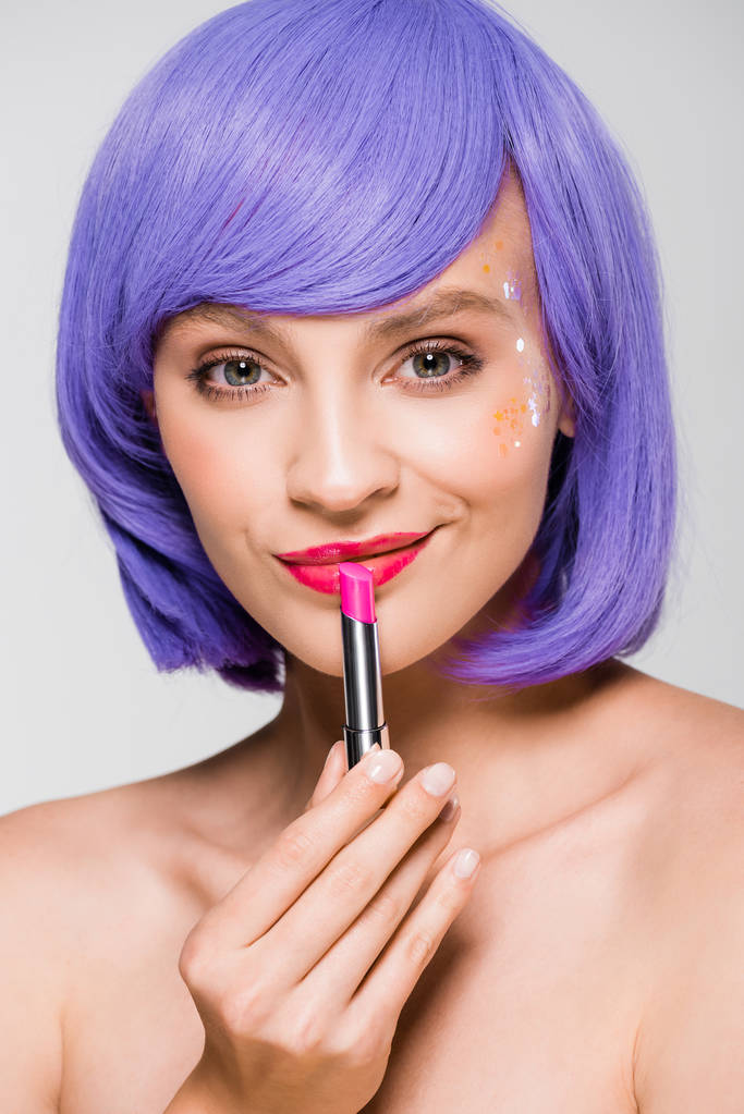 bonita chica en peluca púrpura sosteniendo lápiz labial aislado en gris
 - Foto, imagen
