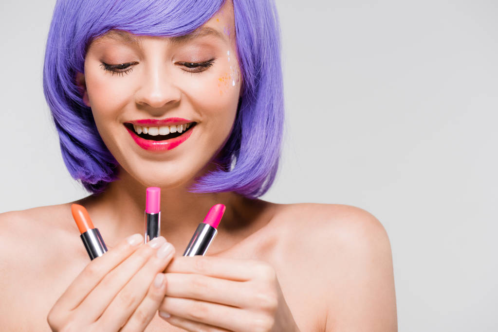 hermosa mujer sonriente en peluca púrpura mirando lápices labiales aislados en gris
 - Foto, Imagen