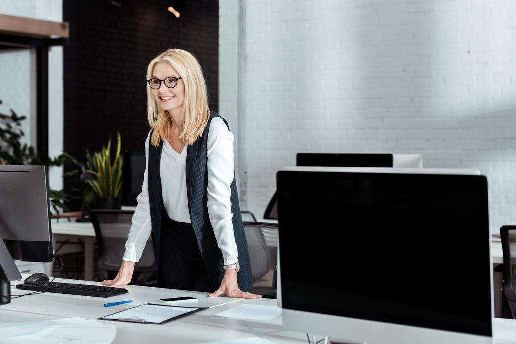 femme d'affaires blonde gaie dans des lunettes debout près du bureau dans le bureau
  - Photo, image