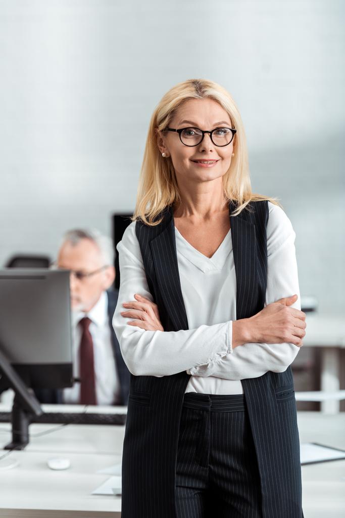 enfoque selectivo de alegre mujer de negocios rubia en gafas de pie con los brazos cruzados cerca de compañero de trabajo
  - Foto, Imagen
