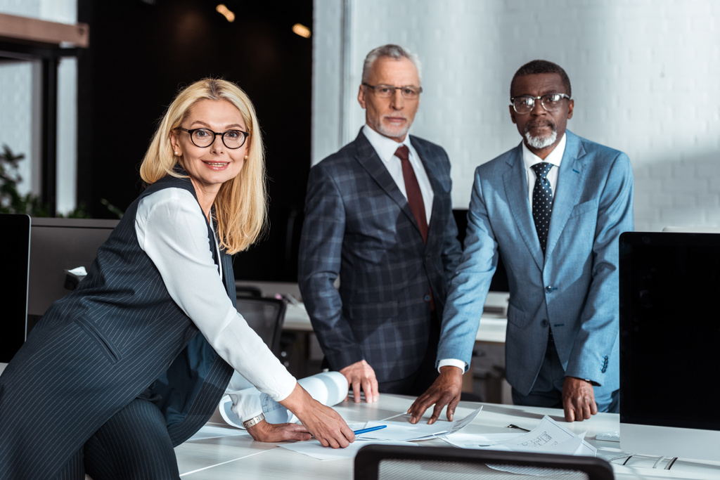 enfoque selectivo de mujer de negocios rubia en gafas de pie cerca de la mesa y hombres de negocios multiculturales
  - Foto, Imagen