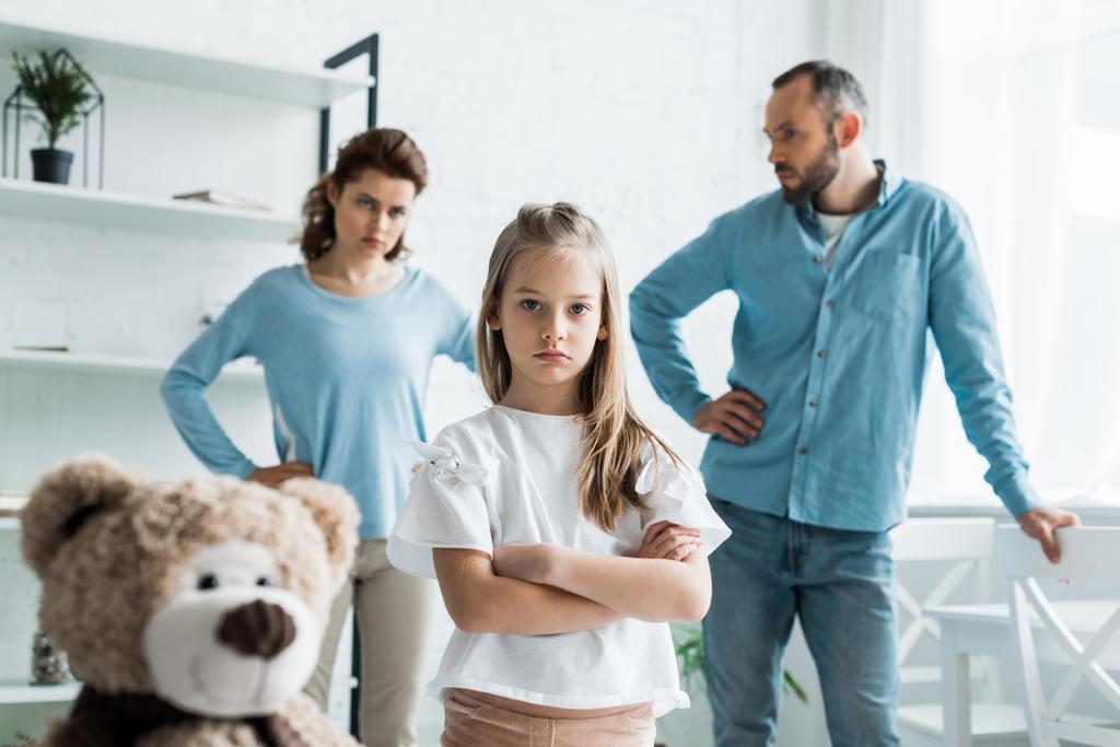selective focus of cute kid standing with crossed arms near teddy bear and parents at home  - Photo, Image