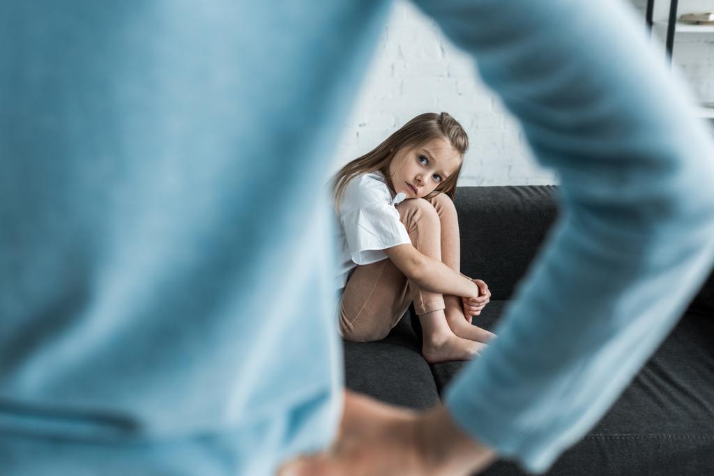 enfoque selectivo del niño asustado sentado en el sofá mirando a la madre de pie con la mano en la cadera
 - Foto, Imagen