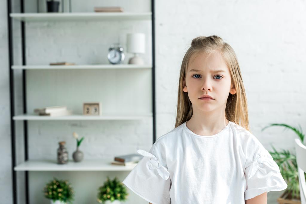 αναστατωμένος παιδί σε λευκό μπλουζάκι κοιτάζοντας την κάμερα στο σπίτι  - Φωτογραφία, εικόνα