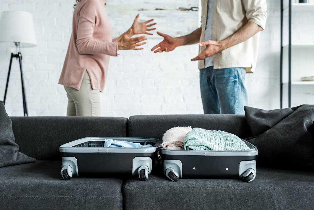 περικομμένη θέα του άντρα και της γυναίκας που στέκονται και την εμφάνιση κοντά σε βαλίτσα με ρούχα στον καναπέ  - Φωτογραφία, εικόνα