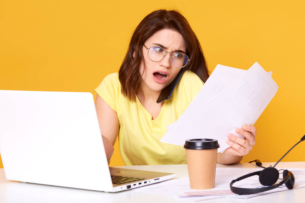 Фото напряженной молодой брюнетки деловая женщина, работающая с бумажными документами в офисе изолированы на желтом фоне, проверка отчетов и найти много ошибок, удивило выражение
. - Фото, изображение