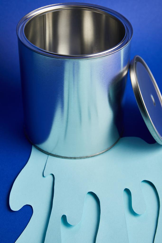 lege metalen glanzend kan en druipend papier knippen verf op heldere blauwe achtergrond - Foto, afbeelding