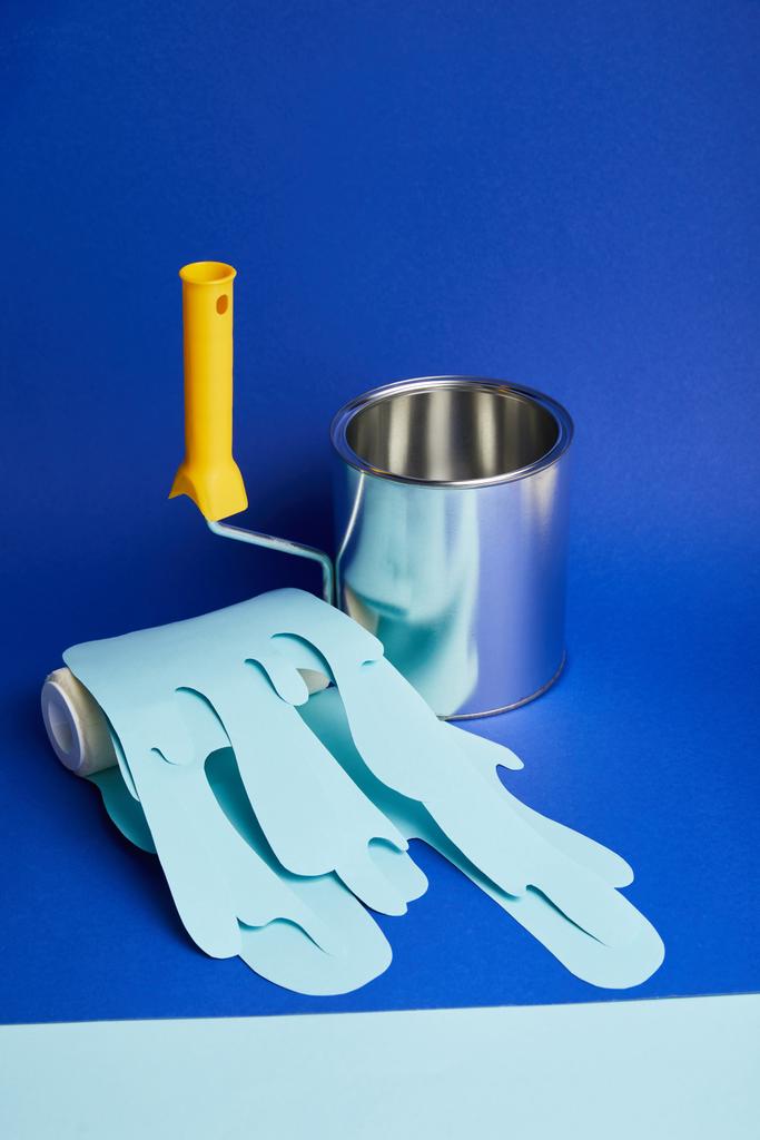 leeg metaal glanzend kan in de buurt van roller met druipend papier knippen verf op heldere blauwe achtergrond - Foto, afbeelding