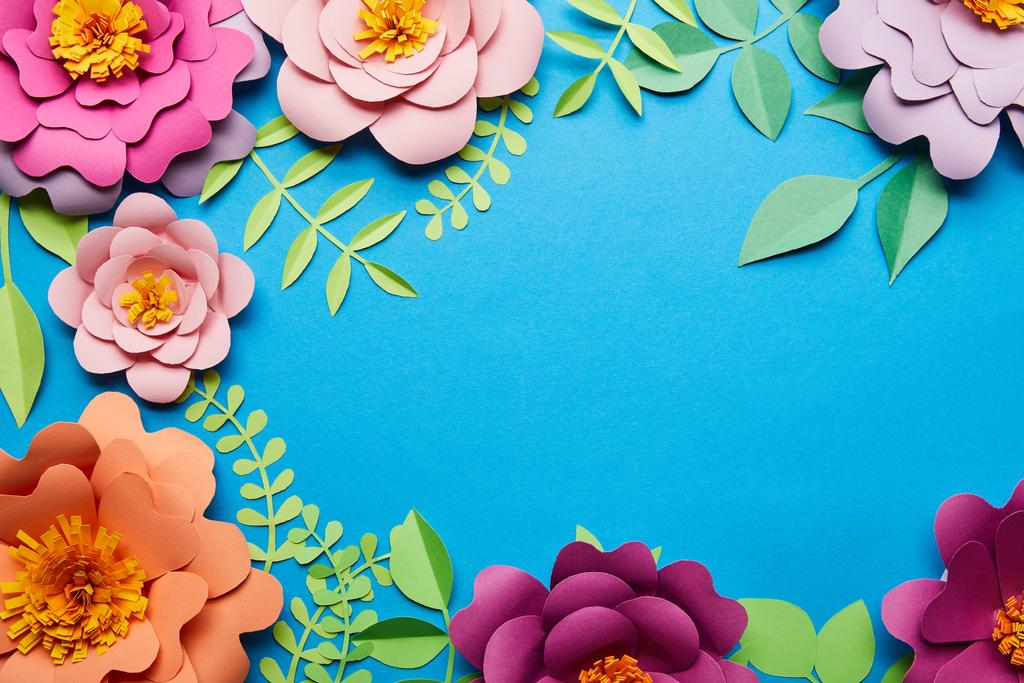 top view of multicolored bright paper cut flowers with green leaves on blue background with copy space - Photo, Image