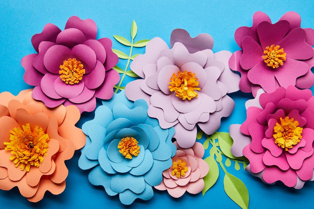 top view of colorful paper cut flowers with green leaves on blue background - Photo, Image