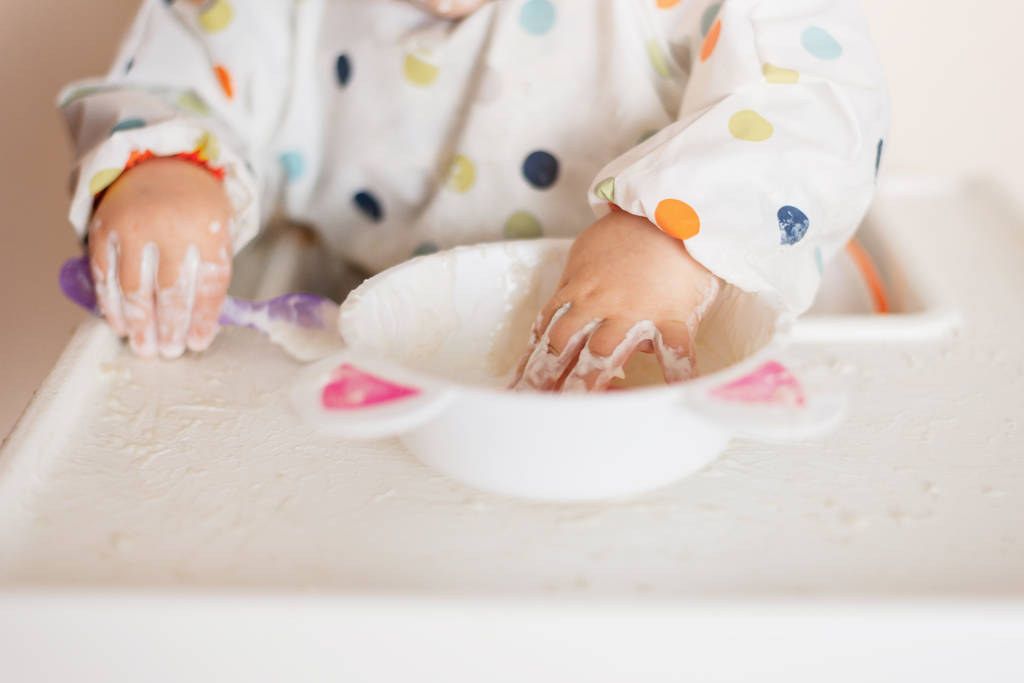 uma menina em uma camiseta e prato sentado em uma cadeira infantil comendo com as mãos cereais com iogurte
 - Foto, Imagem