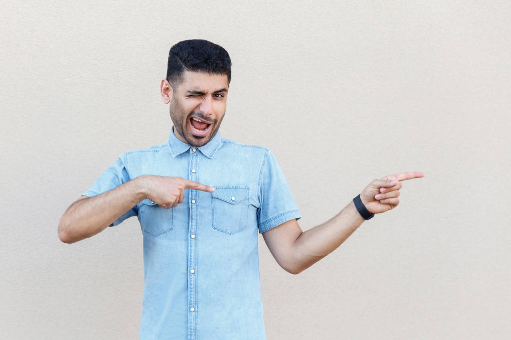 funny handsome young bearded man in blue shirt making crazy face and winking while looking at camera and pointing aside on light beige wall background - Photo, Image
