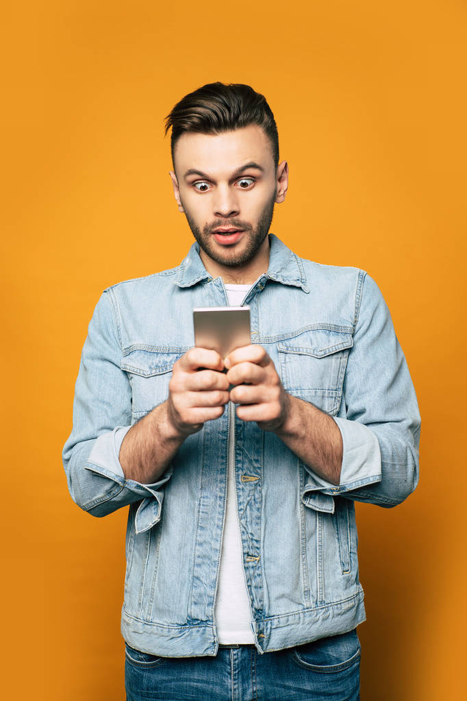 junger moderner stylischer Mann in Jeans benutzt Smartphone in den Händen, während er über gelbem Hintergrund steht - Foto, Bild
