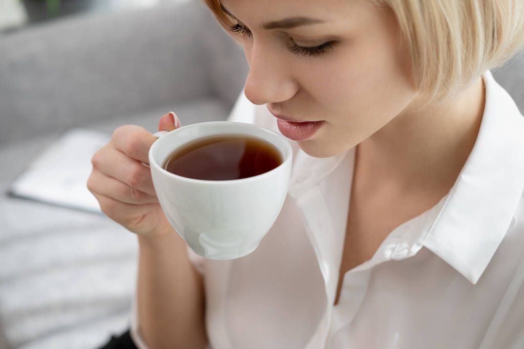 Young beautiful blonde girl with short hair in a white shirt and glasses is sitting on the sofa in bright in the office against the window. Holds a white cup and saucer. Drinking coffee or tea in a - Photo, Image