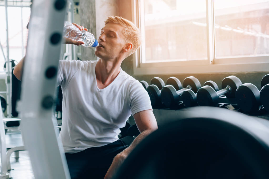 Młody biały mężczyzna na siłowni, siedzi na ławce i pije wodę z butelki. Pragnienie i odwodnienie - Zdjęcie, obraz
