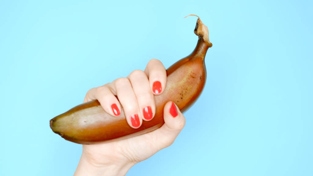 γυναικεία χέρια με κόκκινα νύχια κρατούν μια κόκκινη μπανάνα σε μπλε φόντο - Φωτογραφία, εικόνα