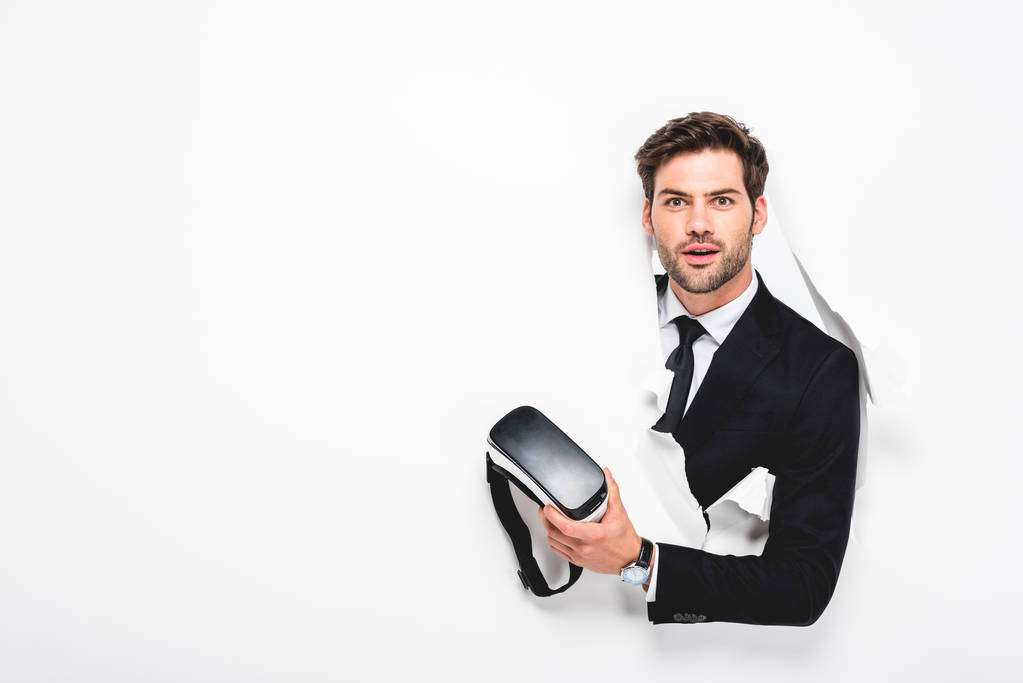 překvapený muž držící sluchátka virtuální realita za dírou ve zdi na bílém - Fotografie, Obrázek
