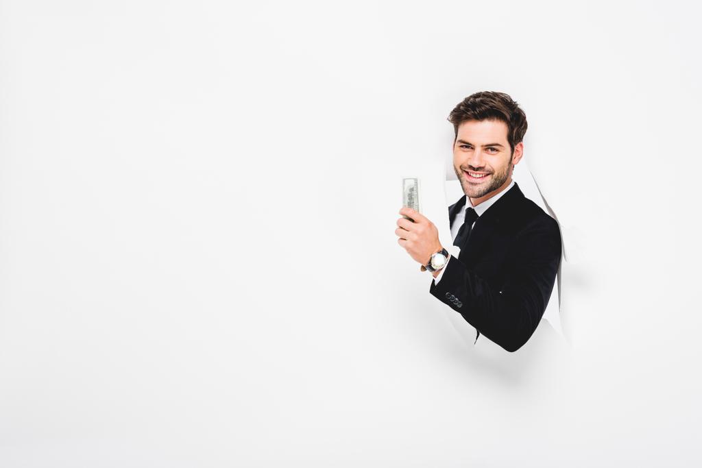 homem de negócios sorrindo segurando notas de dólar atrás do buraco na parede de papel em branco
 - Foto, Imagem