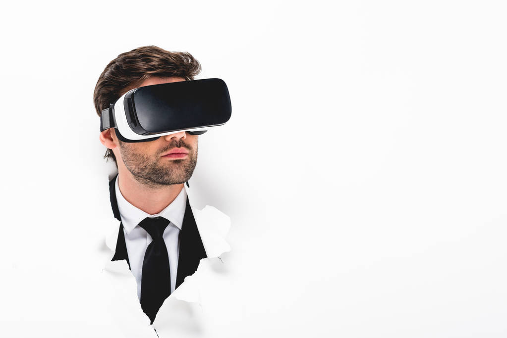 άντρας σε τυπική φθορά και ακουστικά εικονικής πραγματικότητας πίσω από την τρύπα σε χαρτί τοίχου σε λευκό με χώρο αντιγραφής - Φωτογραφία, εικόνα