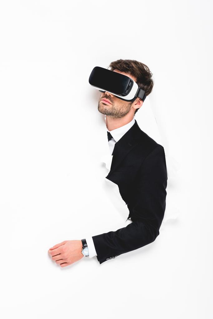 άντρας σε τυπική φθορά και ακουστικά εικονικής πραγματικότητας πίσω από την τρύπα σε χαρτί τοίχου σε λευκό - Φωτογραφία, εικόνα
