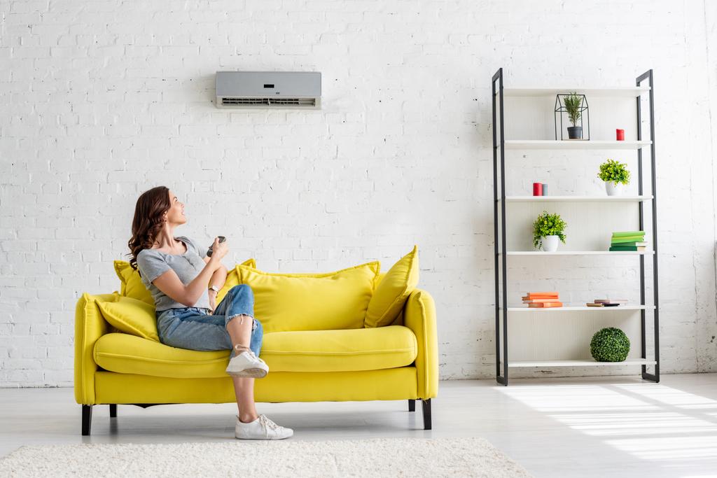 hübsche junge Frau sitzt zu Hause auf gelbem Sofa unter Klimaanlage - Foto, Bild