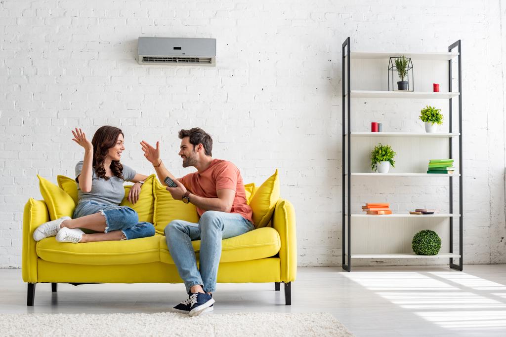 Fröhlicher Mann und Frau im Gespräch auf gelbem Sofa unter Klimaanlage - Foto, Bild