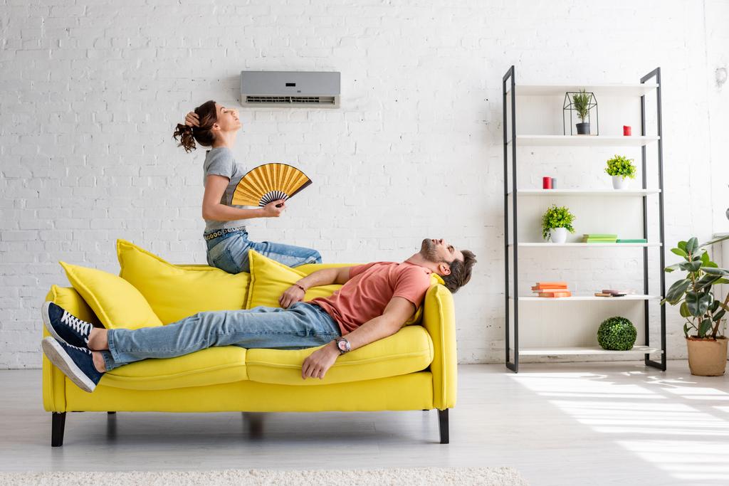 wyczerpane człowiek leżący na żółtej kanapie pod klimatyzator w pobliżu kobiety z wentylatorem dłoni - Zdjęcie, obraz