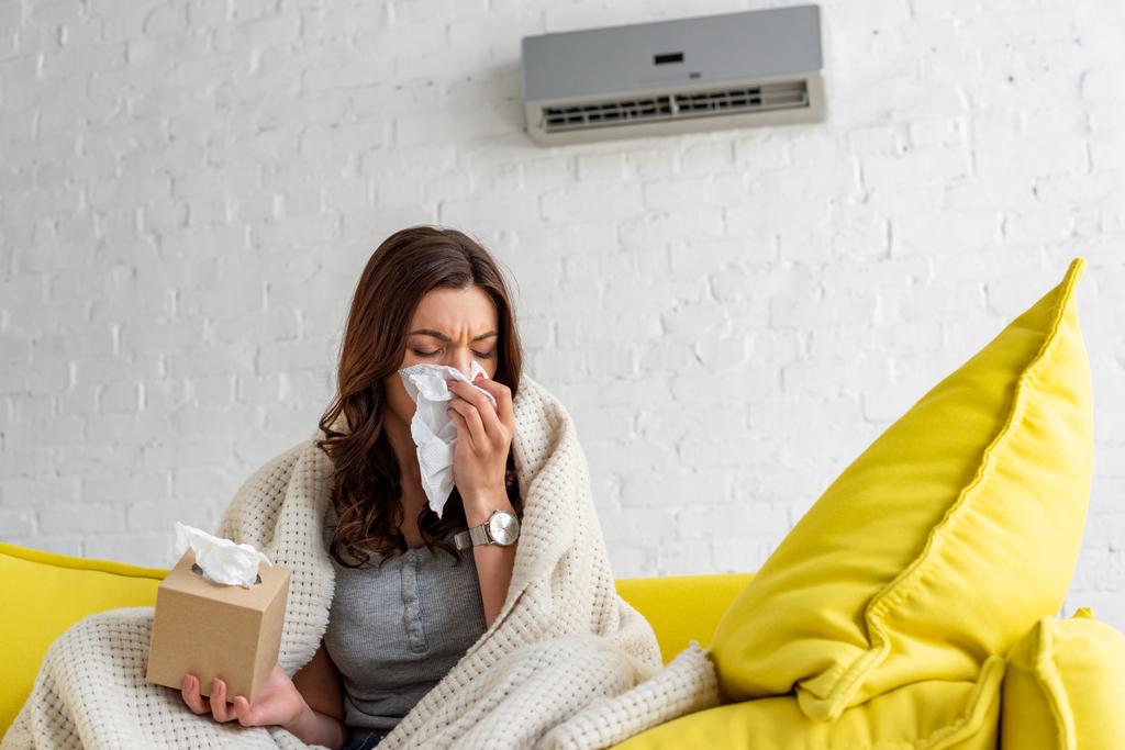 Kranke Frau mit Papierserviette wärmt sich unter Decke, während sie zu Hause unter Klimaanlage sitzt - Foto, Bild