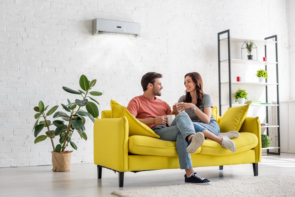 boldog fiatal pár beszél, és gazdaság csészék ülve sárga kanapé alatt légkondicionáló otthon - Fotó, kép