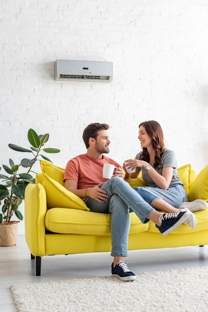 schöner Mann mit hübscher Freundin, die Tassen hält, während sie zu Hause auf dem Sofa unter der Klimaanlage sitzt - Foto, Bild