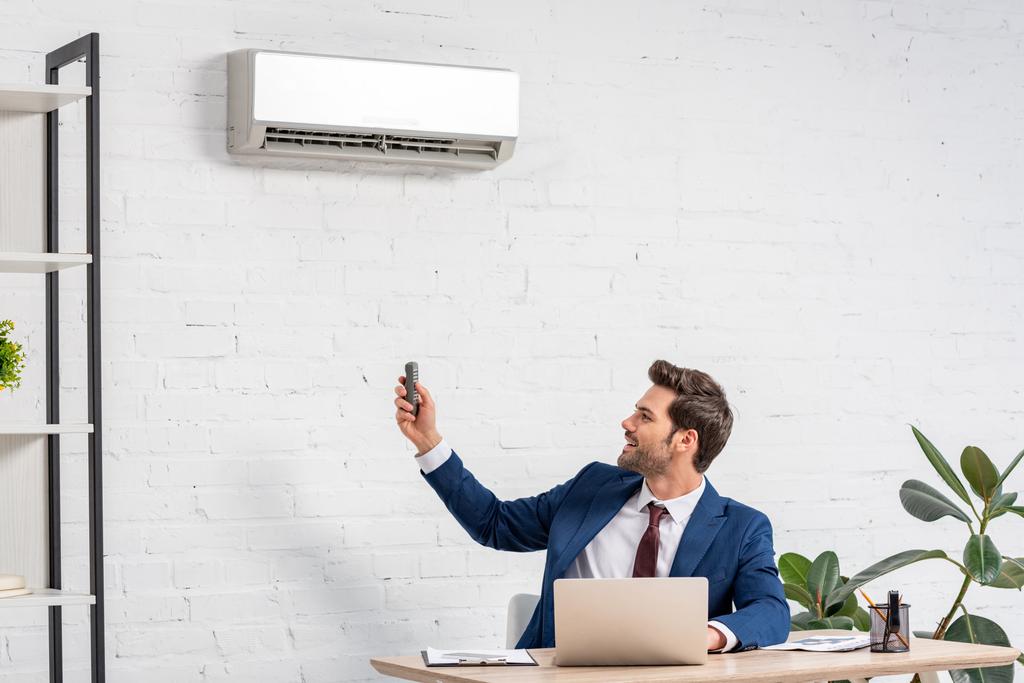 handsome businessman holding remote control while sitting at workplace under air conditioner - Photo, Image