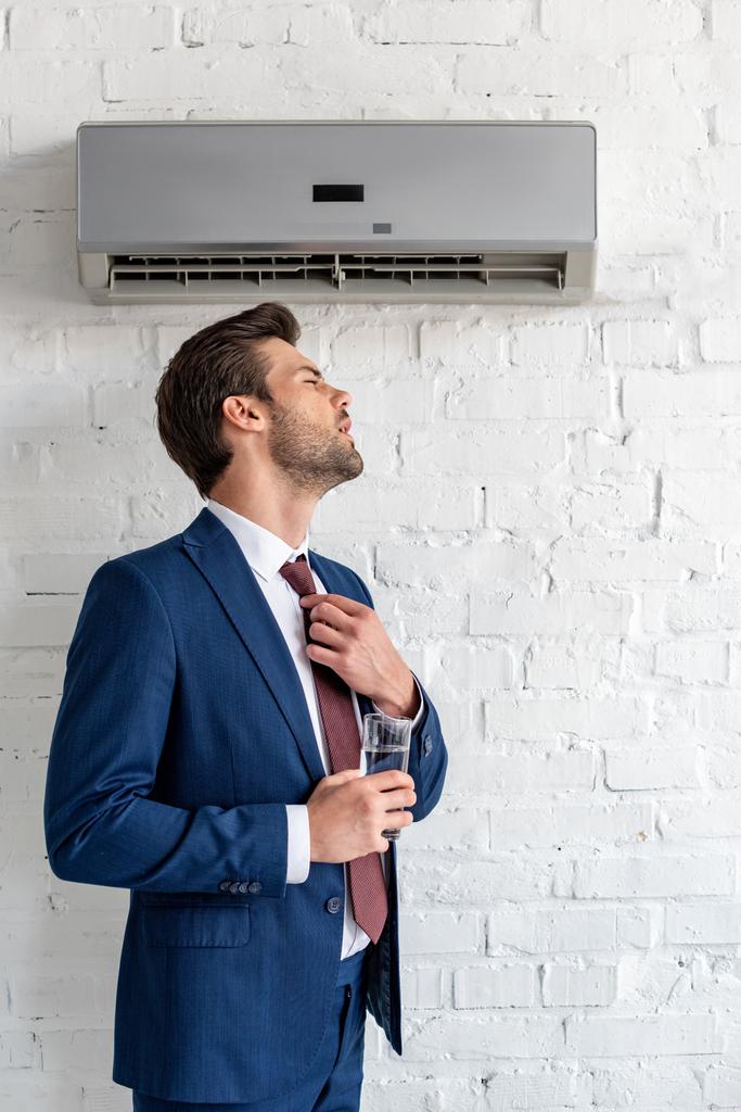 schöner Geschäftsmann mit einem Glas Wasser im Stehen unter einer Klimaanlage - Foto, Bild