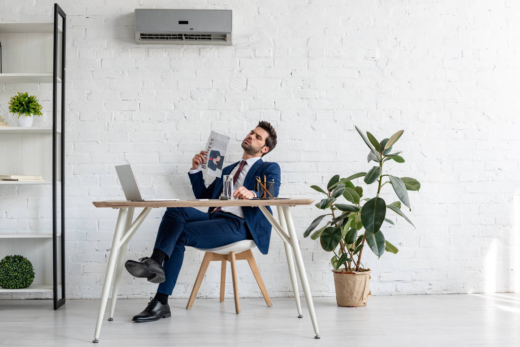 handsome businessman waving business newspaper while sitting at workplace and suffering from heat in office - Photo, Image