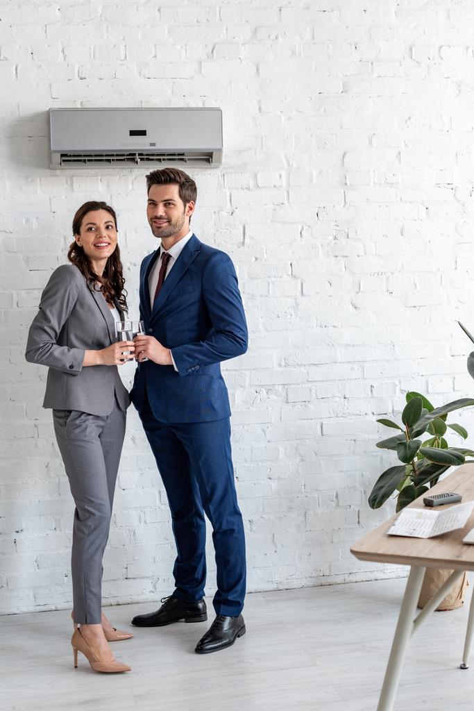 усміхнені бізнесмени тримають склянки води, стоячи під кондиціонером в офісі
 - Фото, зображення