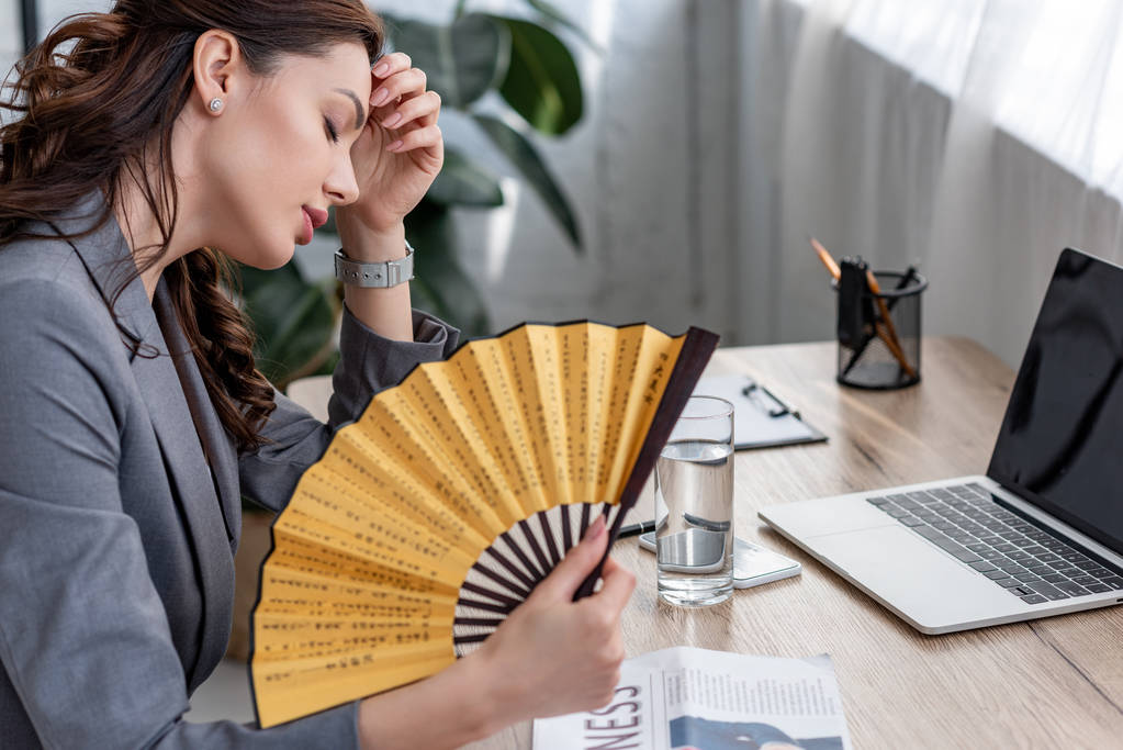 молодая деловая женщина, размахивающая ручным вентилятором, сидя на рабочем месте и страдая от жары в офисе
 - Фото, изображение