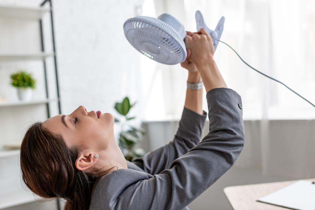 mooie zakenvrouw met gesloten ogen die blaas ventilator vasthouden terwijl ze aan warmte in het kantoor lijden - Foto, afbeelding