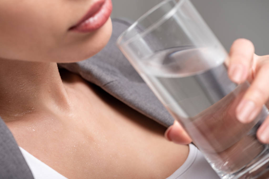 Ausgeschnittene Ansicht einer jungen Frau, die ein Glas Wasser in der Hand hält, während sie an Hitze auf grau leidet - Foto, Bild