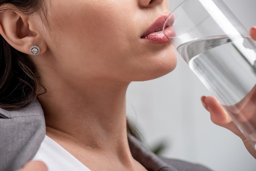 обрезанный вид молодой женщины, страдающей от жары и питьевой воды из стекла на сером
 - Фото, изображение