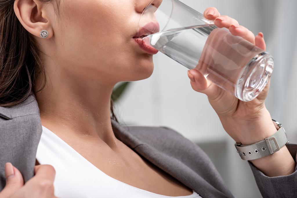 Teilbild einer jungen Geschäftsfrau, die Wasser aus Glas trinkt, während sie unter Hitze leidet - Foto, Bild
