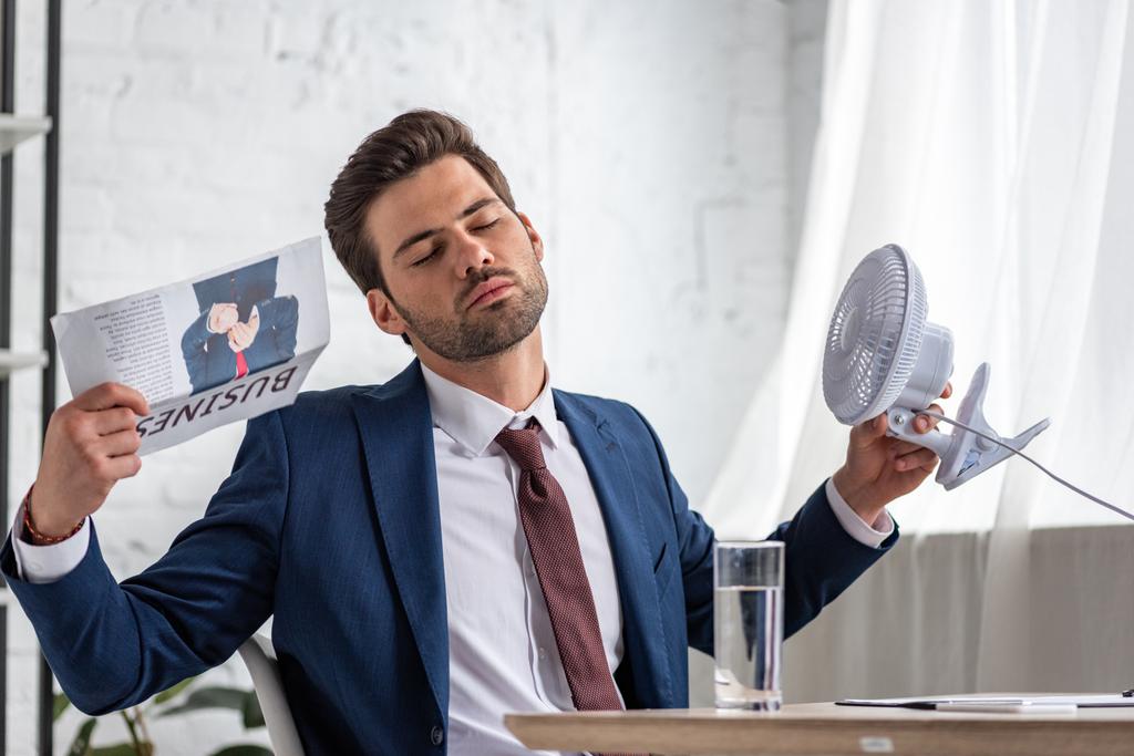 bonito empresária segurando ventilador elétrico e acenando jornal de negócios enquanto sofrendo de calor no escritório
 - Foto, Imagem
