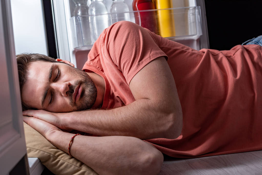 knappe vermoeide man slaapt op de vloer in de keuken in de buurt van open koelkast  - Foto, afbeelding