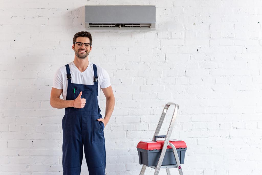 reparador alegre mostrando polegar para cima enquanto está em pé sob ar condicionado perto de escada rolante e caixa de ferramentas
 - Foto, Imagem