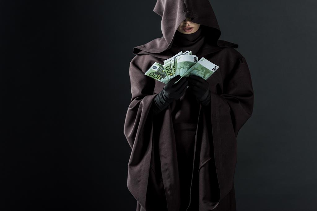 黒で隔離されたユーロ紙幣を保持する死の衣装を着た女性のトリミングされたビュー - 写真・画像
